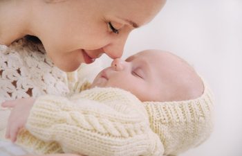 A woman gently touches her nose to a sleeping baby’s nose while holding the baby, both wearing cream-colored knit clothing.